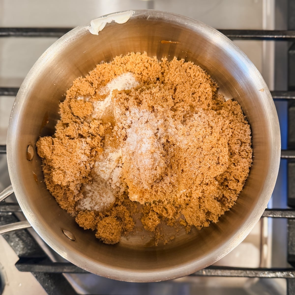 A stainless steel saucepan on a stovetop contains a mixture of brown sugar and butter, ready to be melted or cooked.