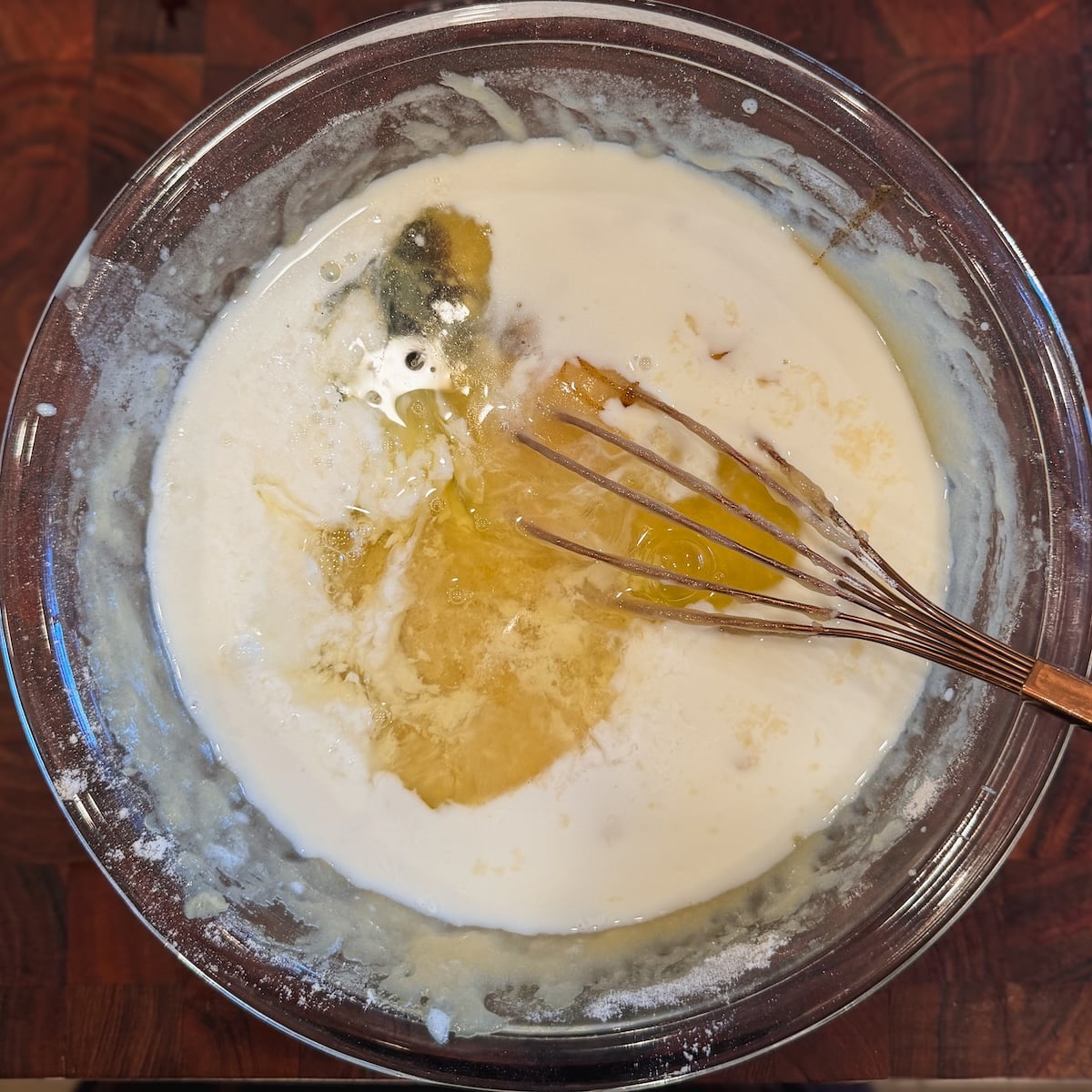 A glass bowl with a whisk mixing wet and dry ingredients, eggs and buttermilk, on a wooden surface.