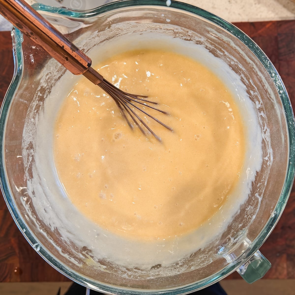 A metal whisk sits in a glass bowl filled with pale yellow cake batter, with some batter clinging to the sides of the bowl on a wooden countertop.