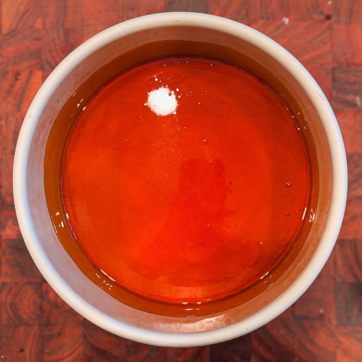 A white bowl filled with bright orange caramel sits on a wooden surface. A small dollop of white substance floats near the edge of the bowl.