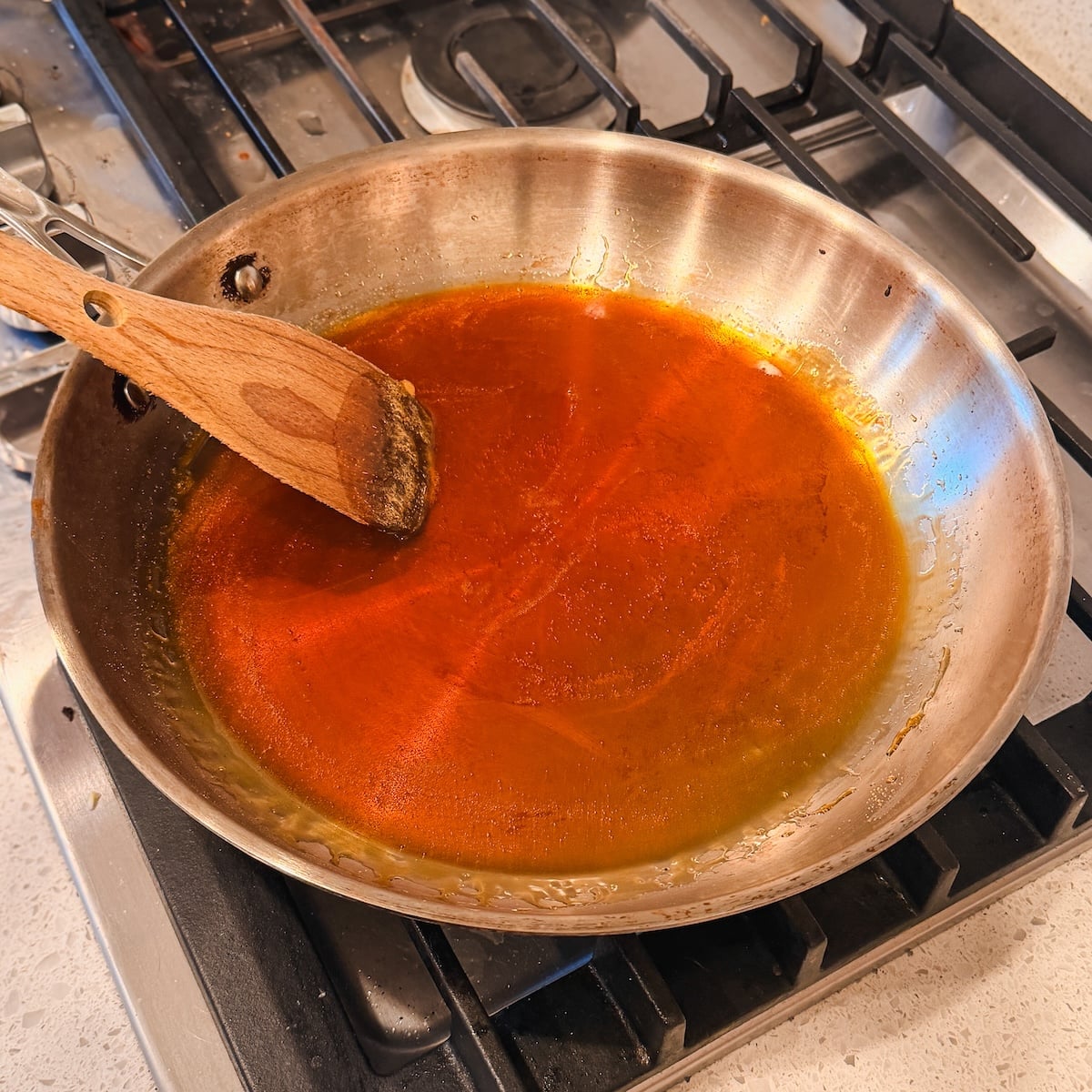 A stainless steel skillet with a wooden spatula rests on a gas stove, containing a layer of golden caramel. The caramel covers the bottom of the pan and appears to be simmering or recently cooked.