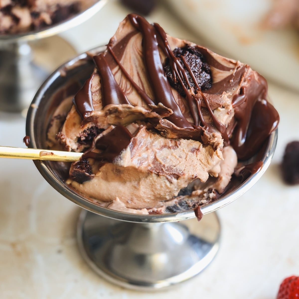 A metal dish filled with chocolate ice cream topped with chocolate sauce and brownie chunks, with a gold spoon resting inside.