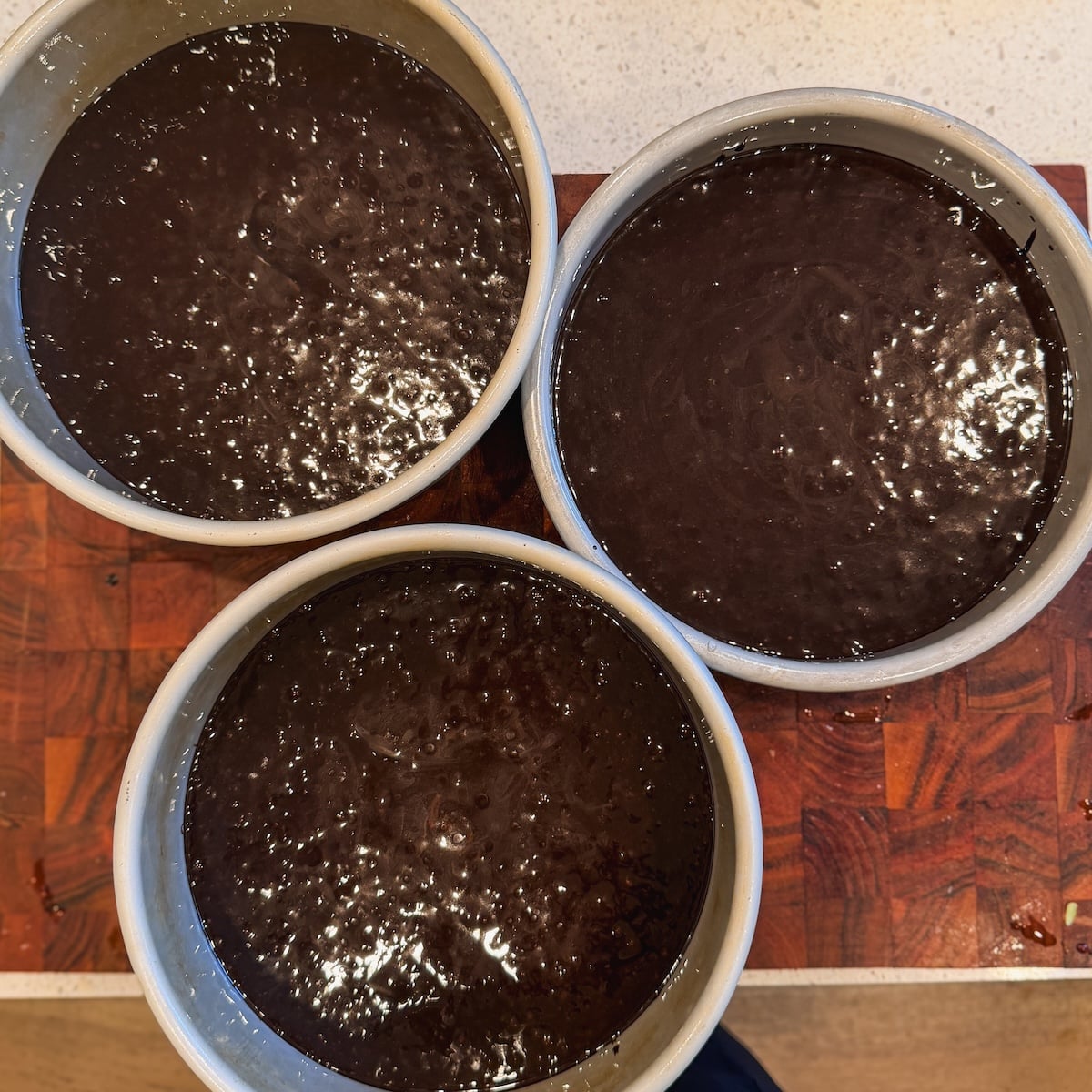 Three round cake pans filled with glossy, dark chocolate cake batter are placed on a wooden surface, ready to be baked.