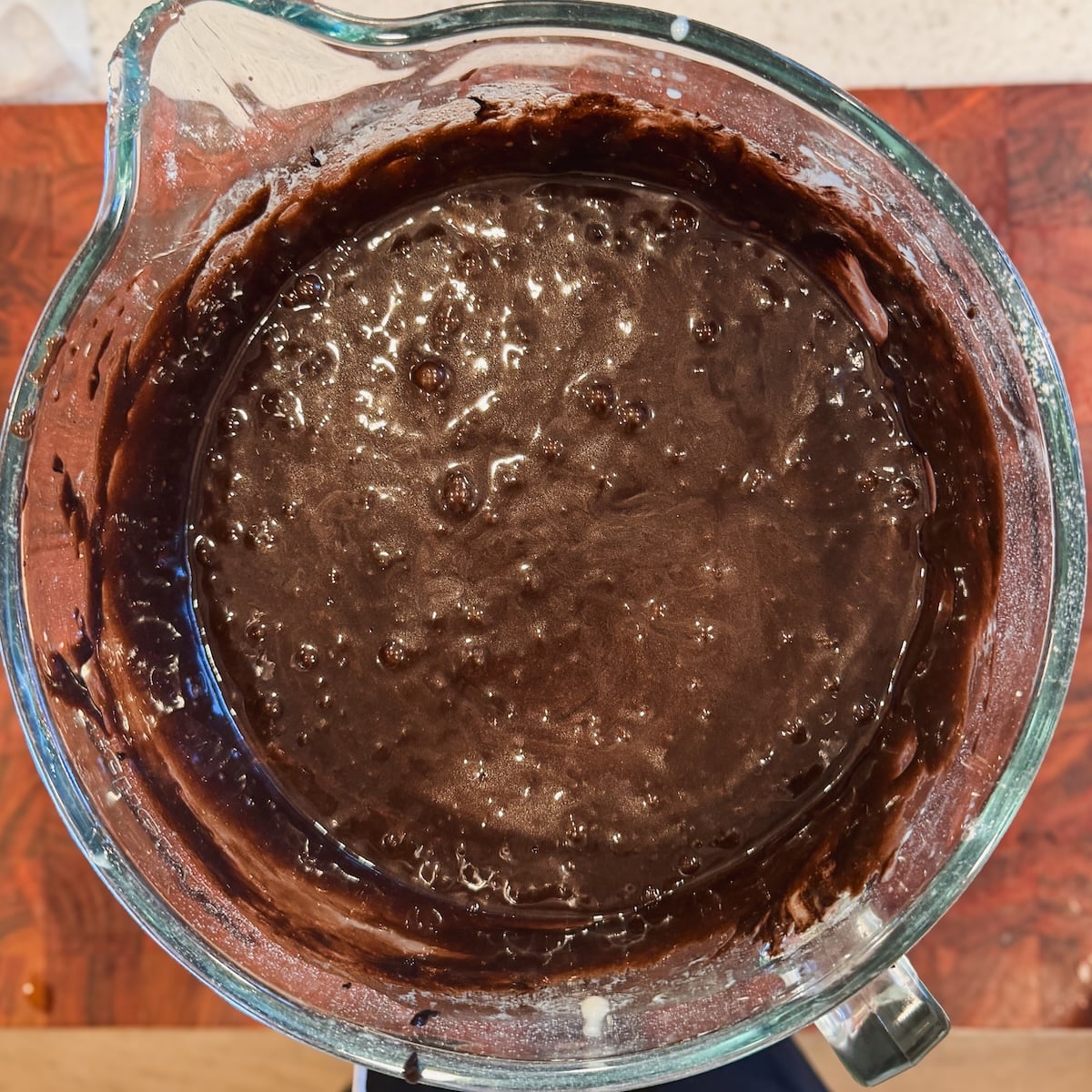 A glass mixing bowl filled with thick, dark chocolate cake batter sits on a wooden countertop.