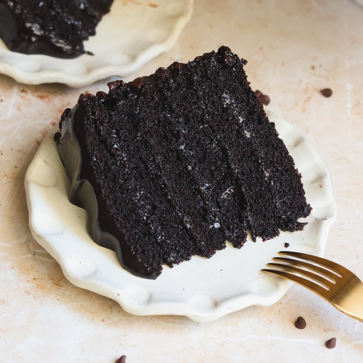 A slice of rich, layered chocolate fudge cake with glossy chocolate icing on a cream-colored plate, next to a gold fork. Chocolate chips are scattered on the light tabletop.