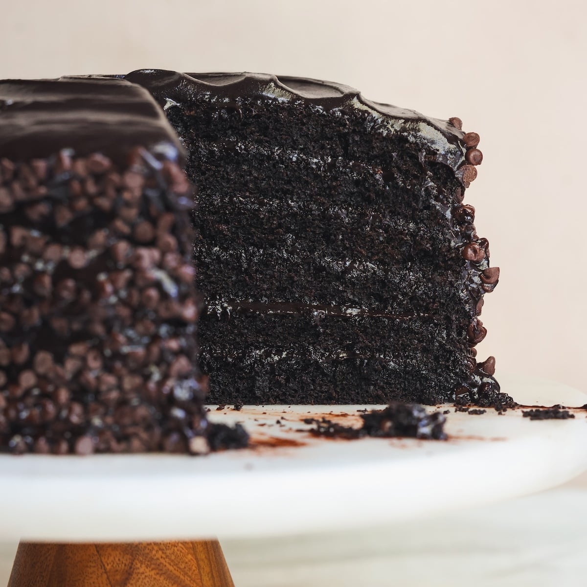 A close-up of a rich, multi-layered chocolate cake with chocolate fudge frosting and chocolate chips, missing one slice, displayed on a white cake stand.
