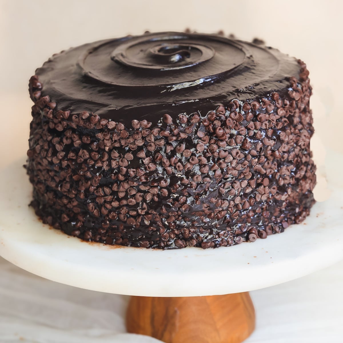 A round chocolate cake covered in glossy chocolate fudge frosting and mini chocolate chips sits on a white marble cake stand with a wooden base.