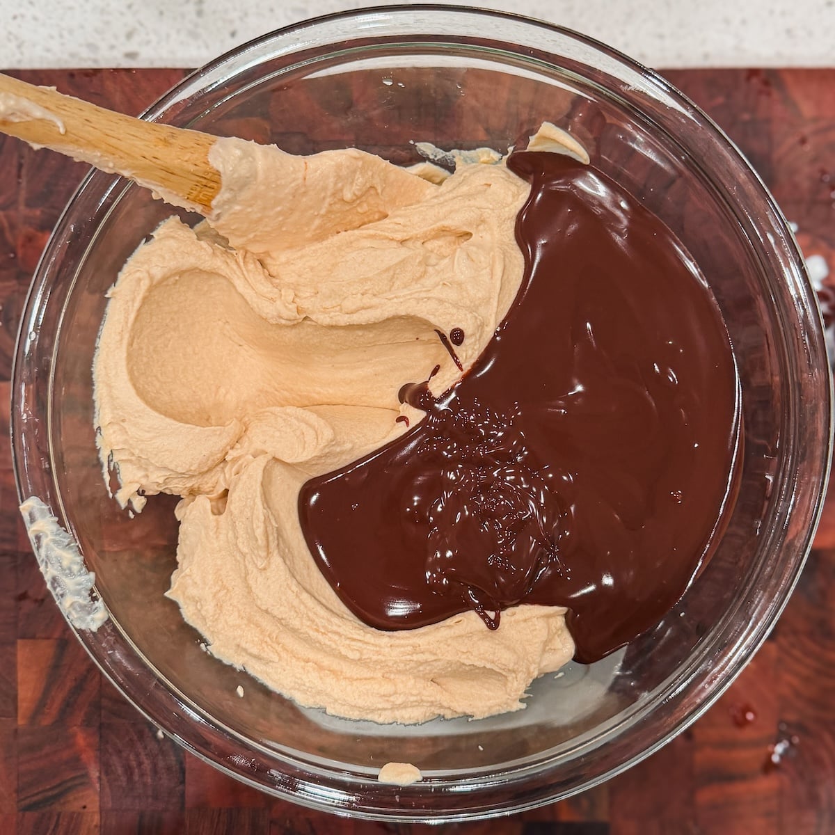 A wooden spatula in a glass bowl filled with an ice cream base partially covered by a layer of chocolate ganache