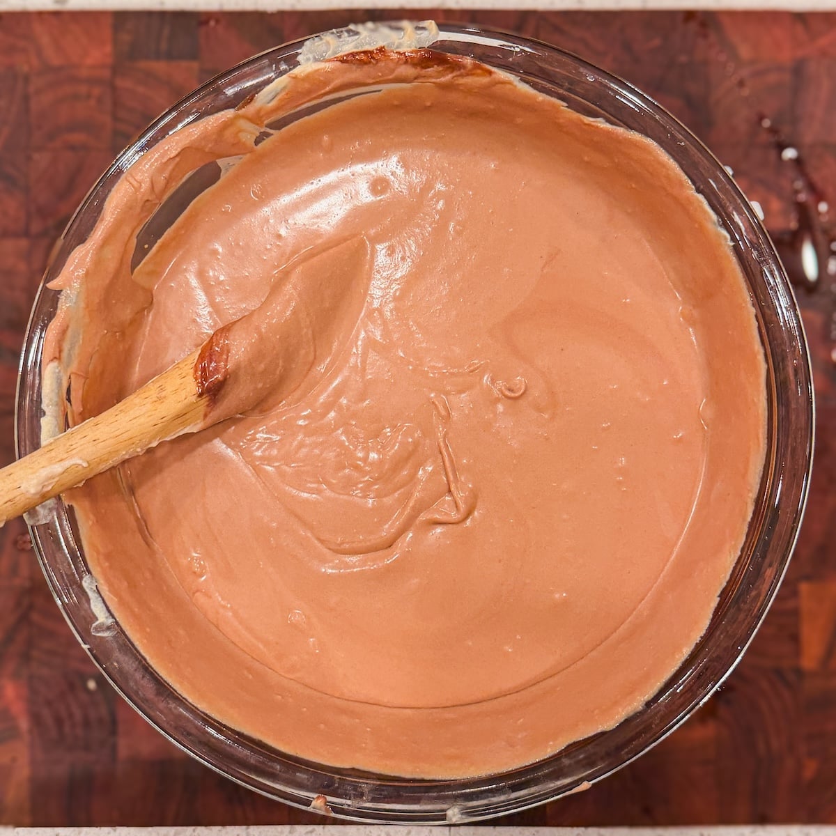 A wooden spatula resting in a glass bowl filled with smooth, light brown chocolate cream mixture