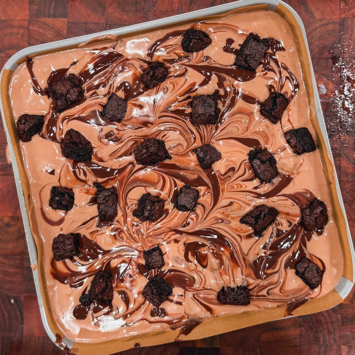 A square pan of chocolate ice cream swirled with ganache and topped with brownie chunks.