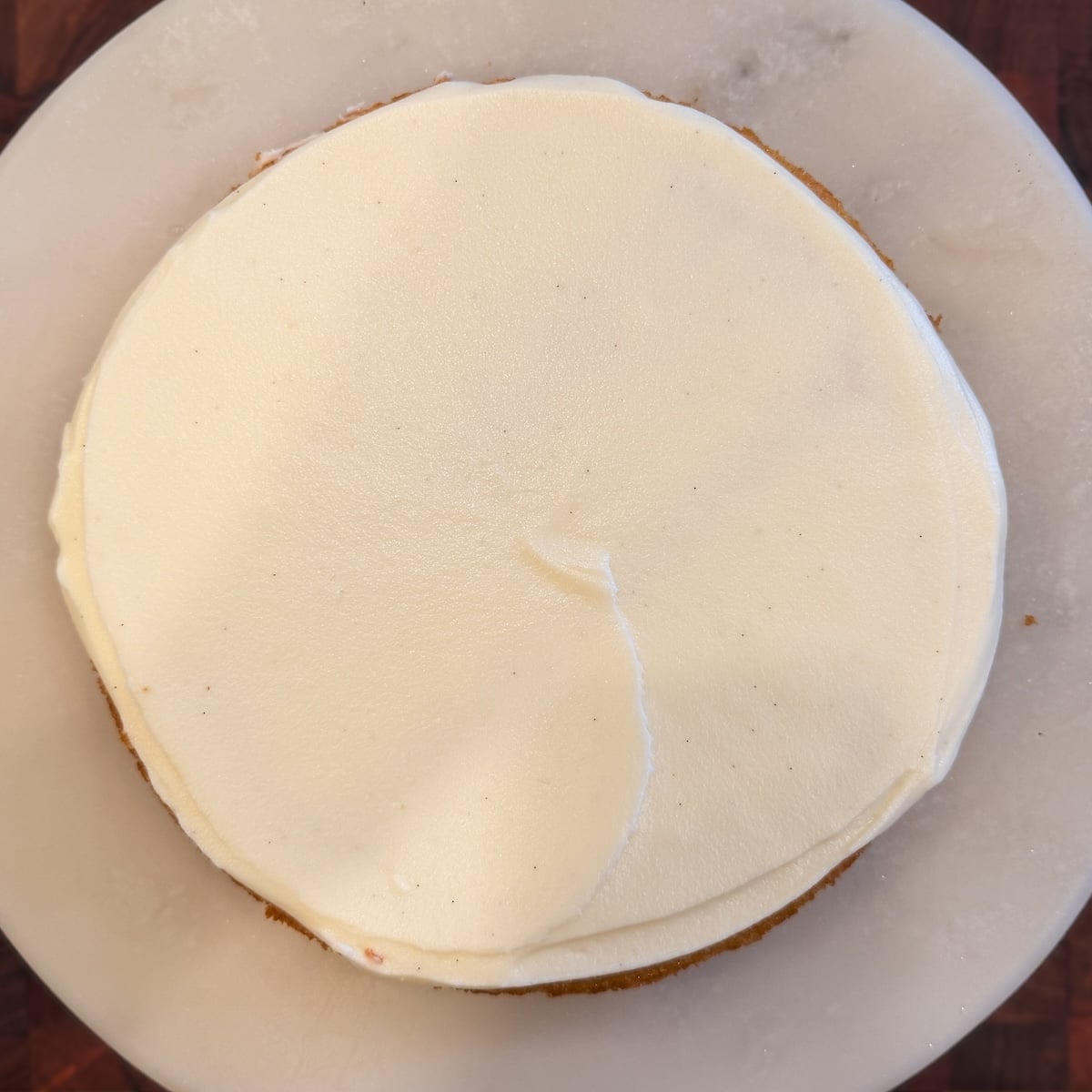 A round cake with smooth cream cheese frosting on top, set on a white marble plate. The frosting has a slight swirl pattern in the center.
