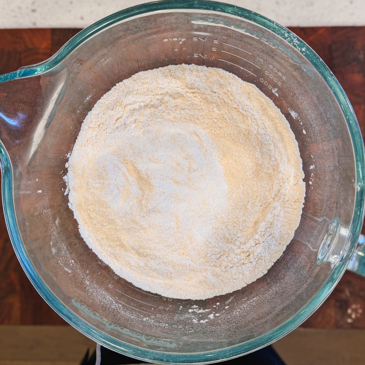 A clear glass mixing bowl filled with a blend of dry ingredients sits on a wooden surface, ready for baking.