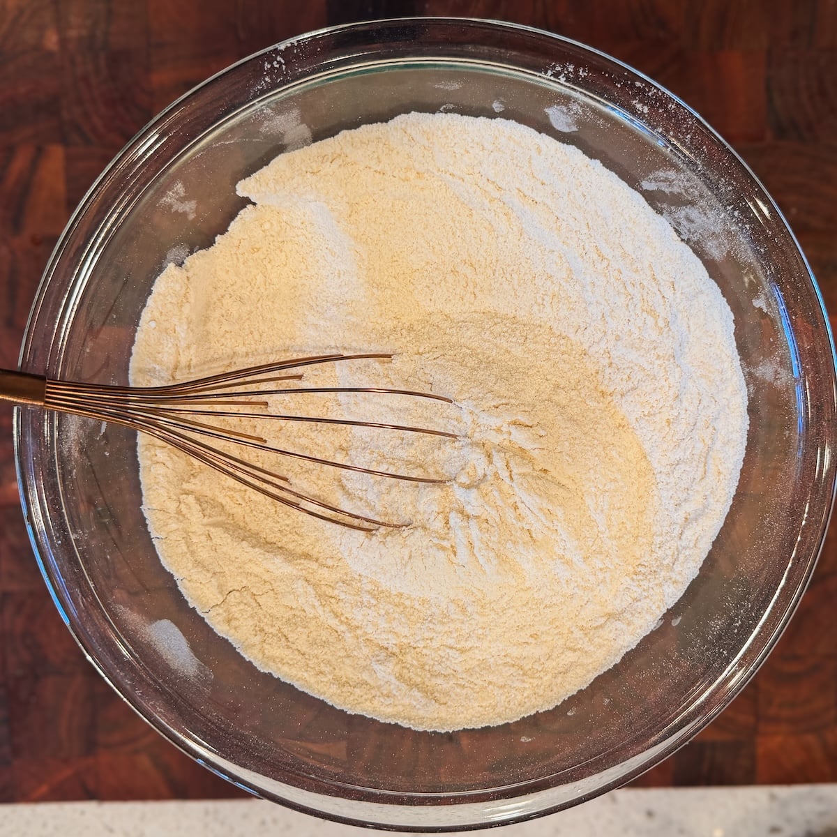 A glass bowl filled with dry ingredients and a wire whisk is resting inside. The bowl sits on a wooden surface