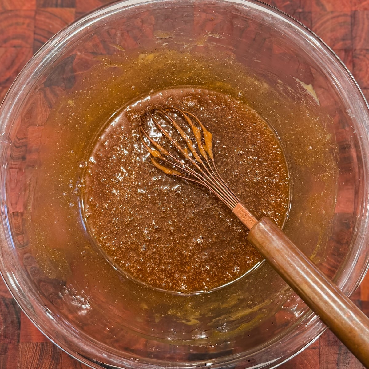A glass bowl with a brown mixture and a whisk resting in it, placed on a wooden surface.