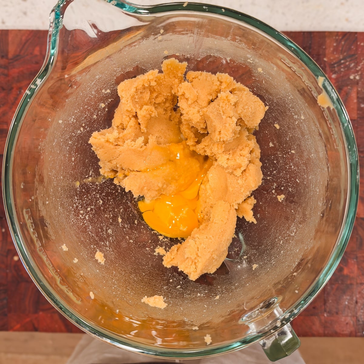 A raw egg and egg yolk cracked into a glass bowl containing a mixture of cookie dough.