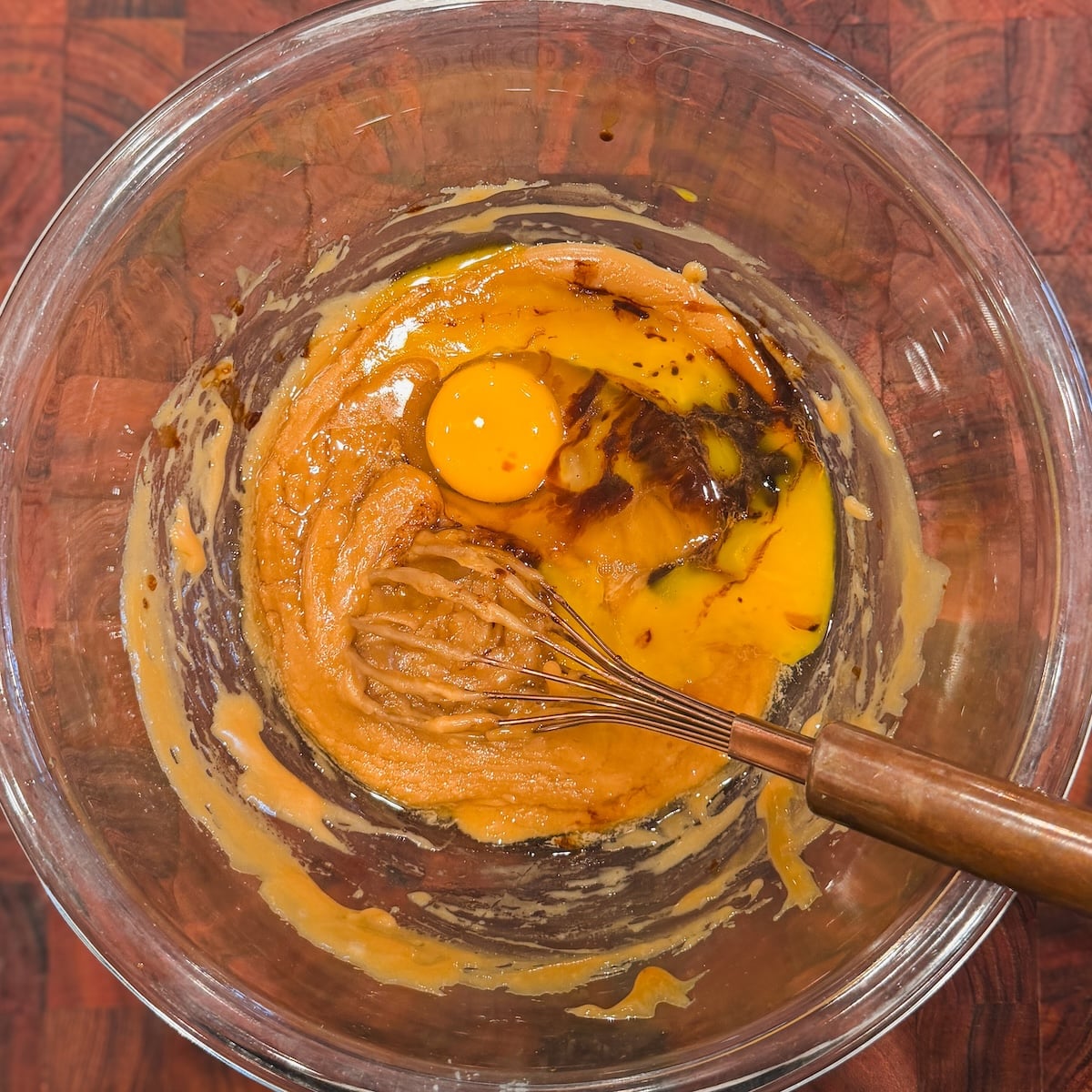A glass bowl with brown sugar, an egg, vanilla, and butter being mixed with a whisk on a wooden surface.