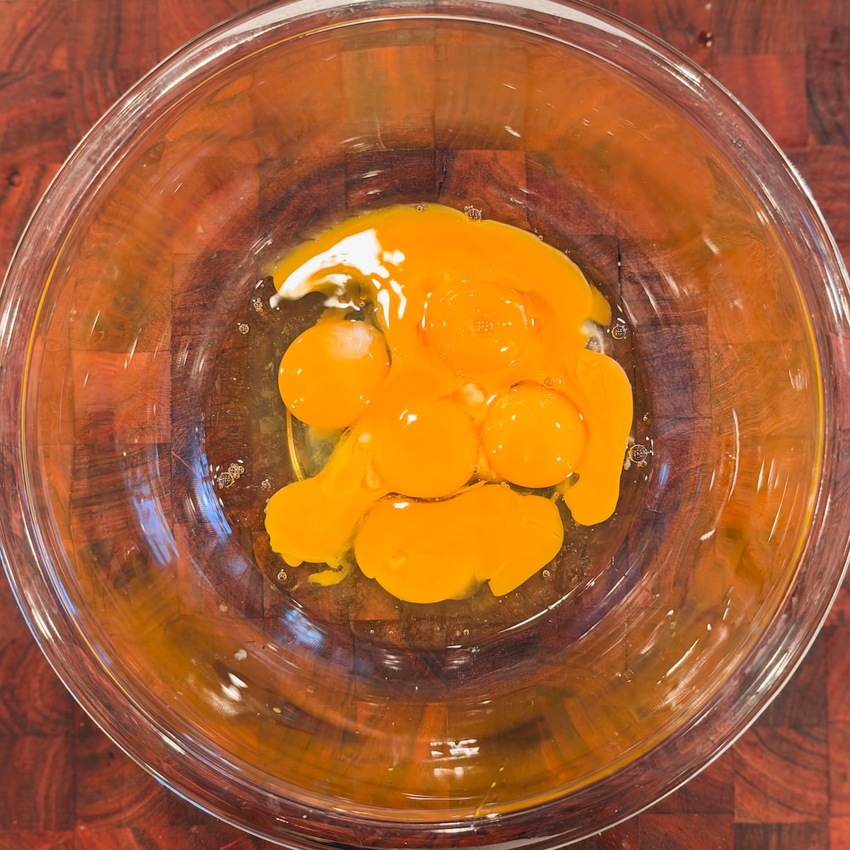 Raw eggs and yolks cracked into a clear glass bowl, sitting on a wooden surface. The yolks and whites are visible and unbroken, ready for mixing or cooking.