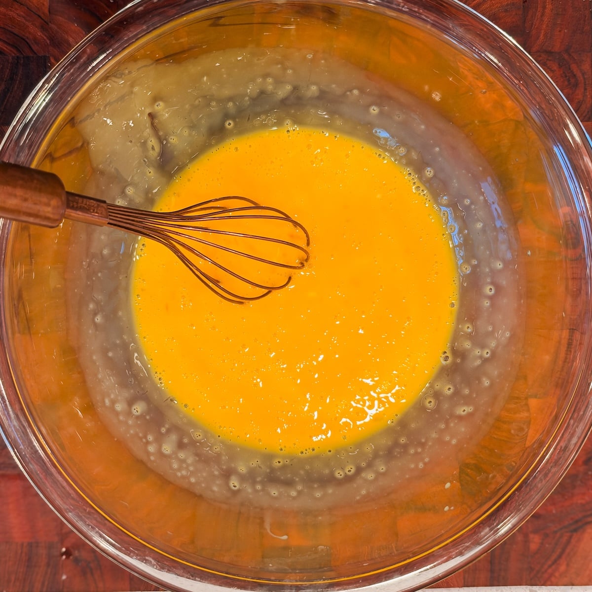 A glass bowl filled with beaten eggs sits on a wooden surface, with a metal whisk resting inside the mixture.