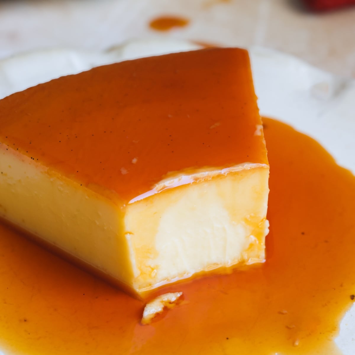 A close-up of a slice of flan topped with golden caramel sauce, served on a white plate. The creamy dessert has a smooth texture, and some caramel sauce pools around its base.