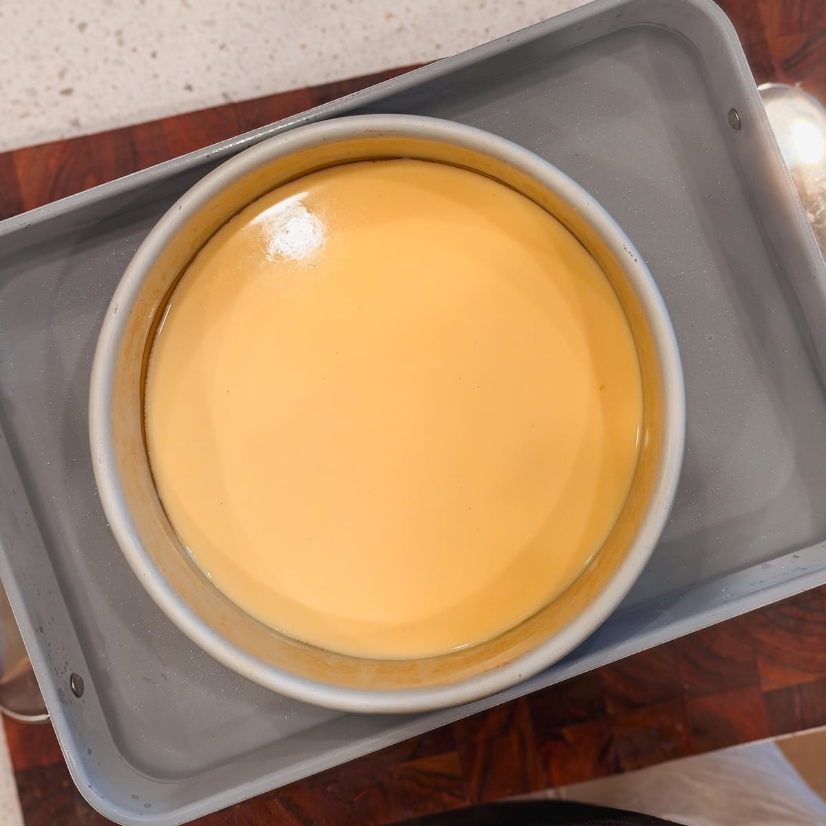 A round pan filled with smooth, yellow custard sits in a larger gray baking dish, likely for a water bath, on a wooden countertop.