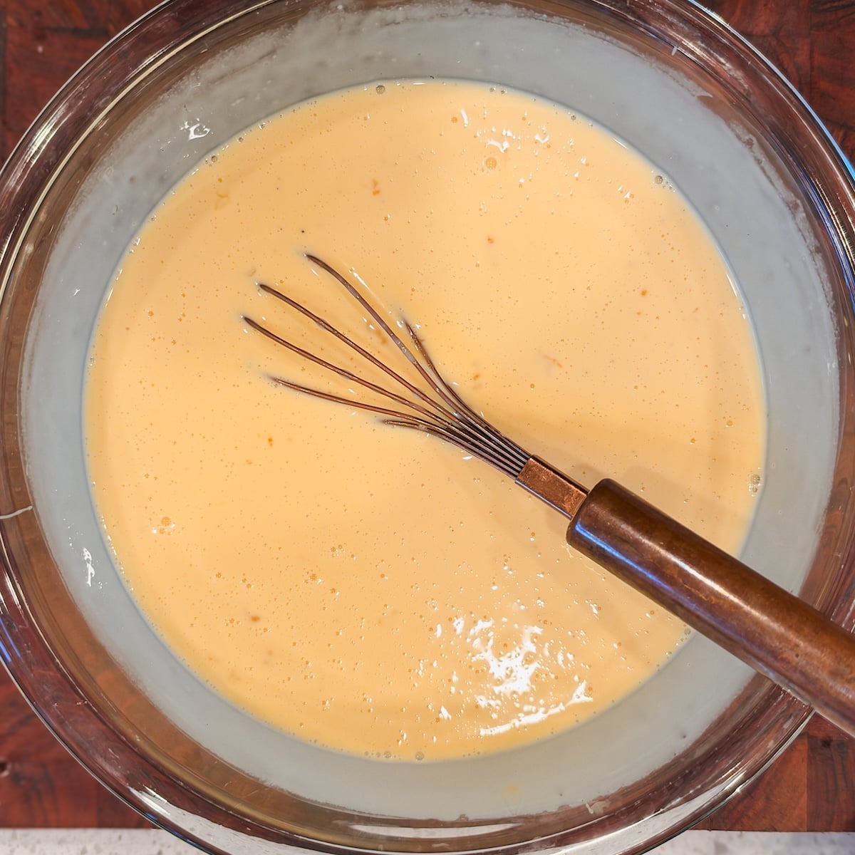 A metal whisk rests in a glass bowl filled with smooth, light yellow custard, sitting on a countertop.