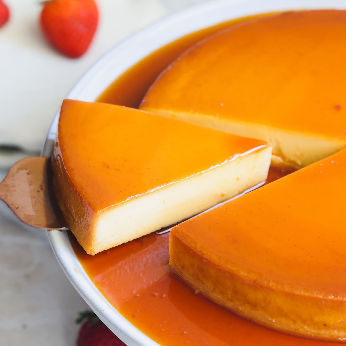 A close-up of a creamy flan in a pan, with one slice being lifted out. The dessert is covered in glossy caramel sauce, and fresh strawberries are visible in the background.