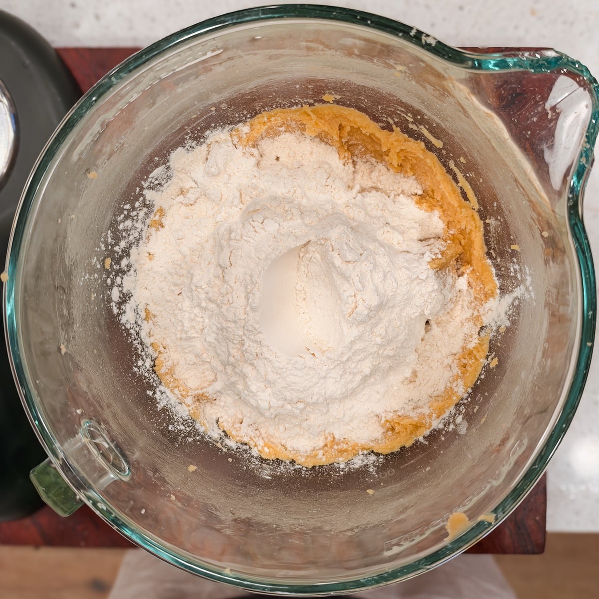 Flour piled on top of cookie dough in a glass stand mixer bowl.