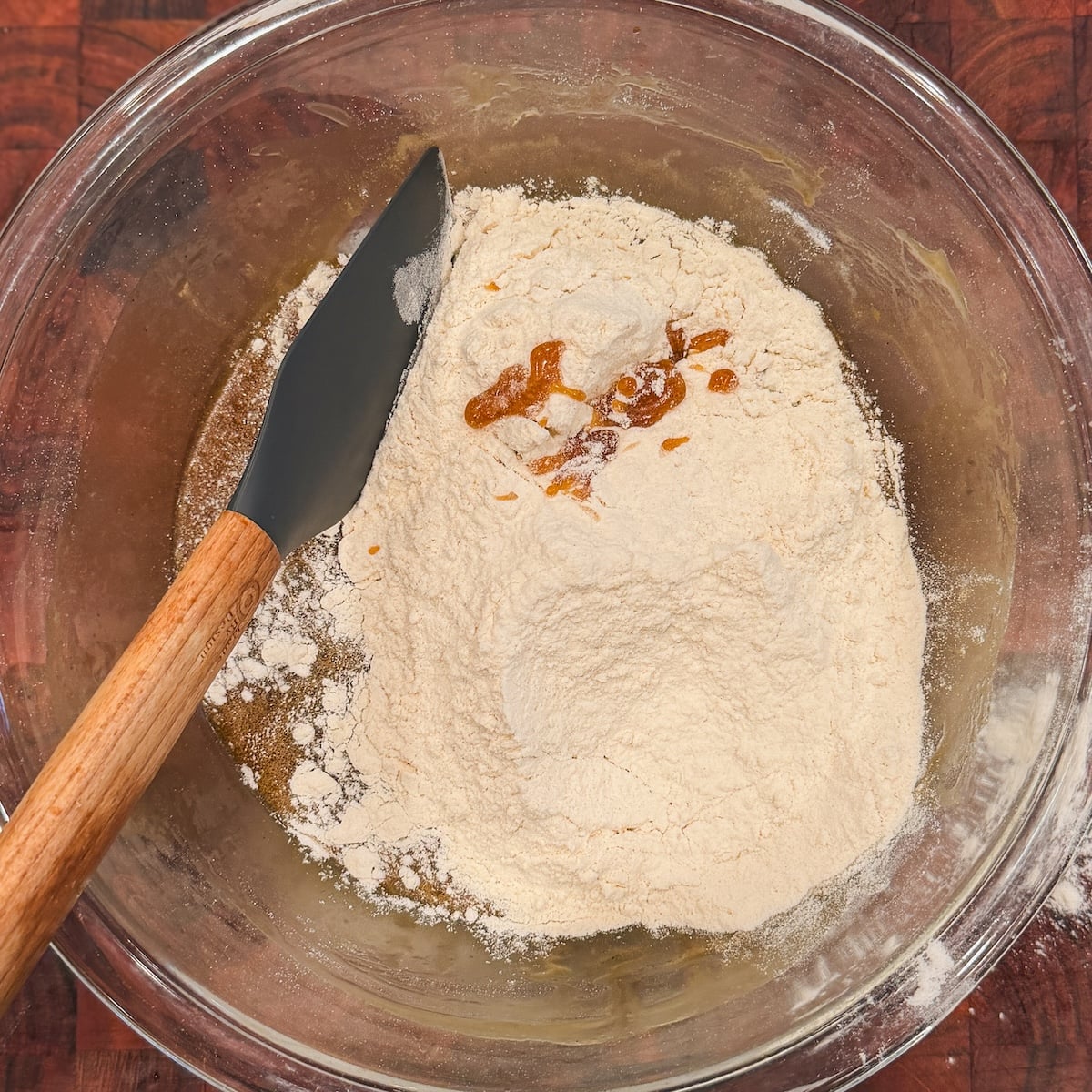 A mixing bowl contains flour, liquid ingredients, and vanilla, about to be mixed with a black spatula with a wooden handle.