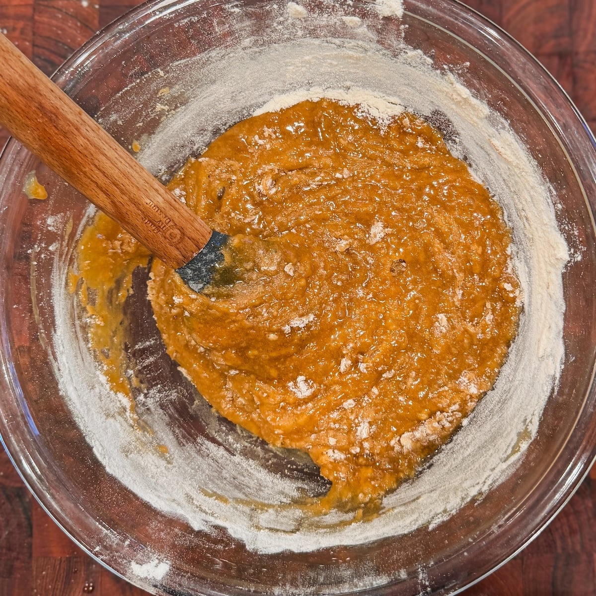 A glass bowl with cookie dough with streaks of flour. The bowl sits on a dark wood surface.