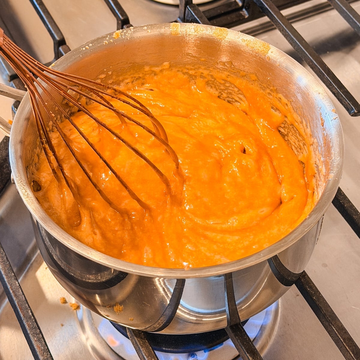 A metal pot on a stove contains thick frothy butterscotch sauce being whisked. The sauce appears to be heating or cooking, as the burner flame is visible beneath the pot.