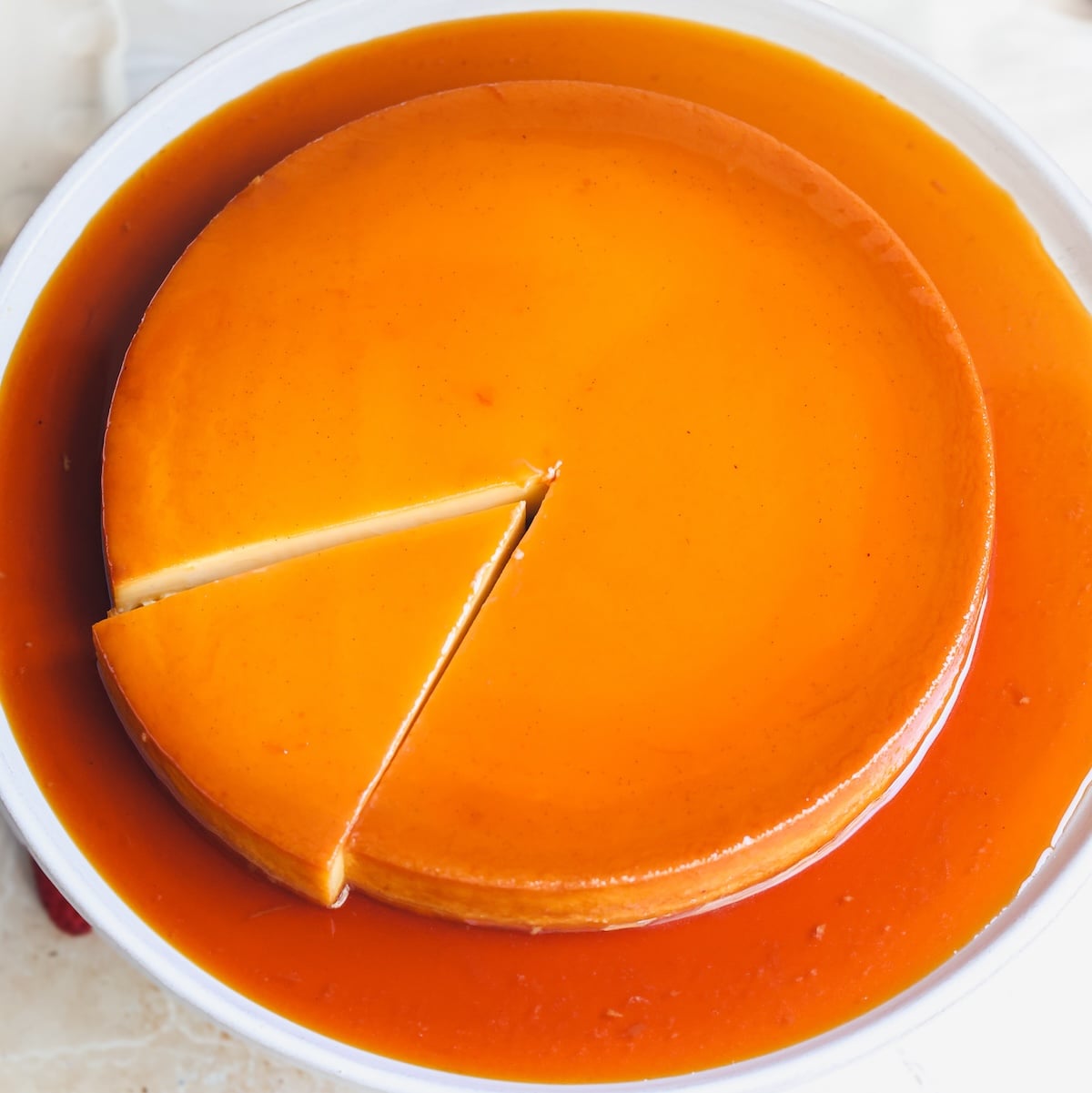 A round flan dessert with caramel sauce sits on a white plate. One slice has been cut and is slightly pulled out from the rest of the flan.