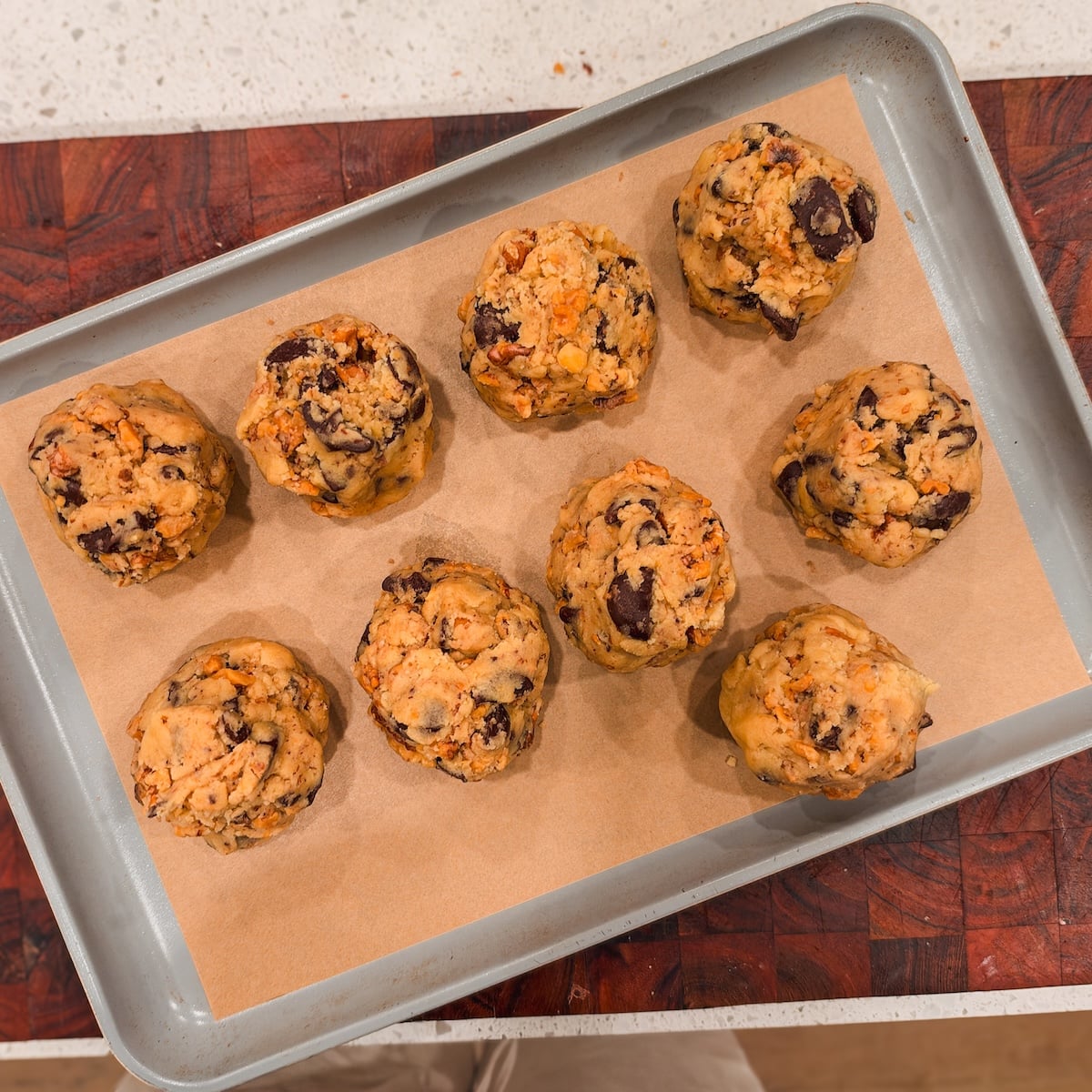 Nine raw big chocolate chip cookie dough balls arranged on a parchment-lined baking sheet.