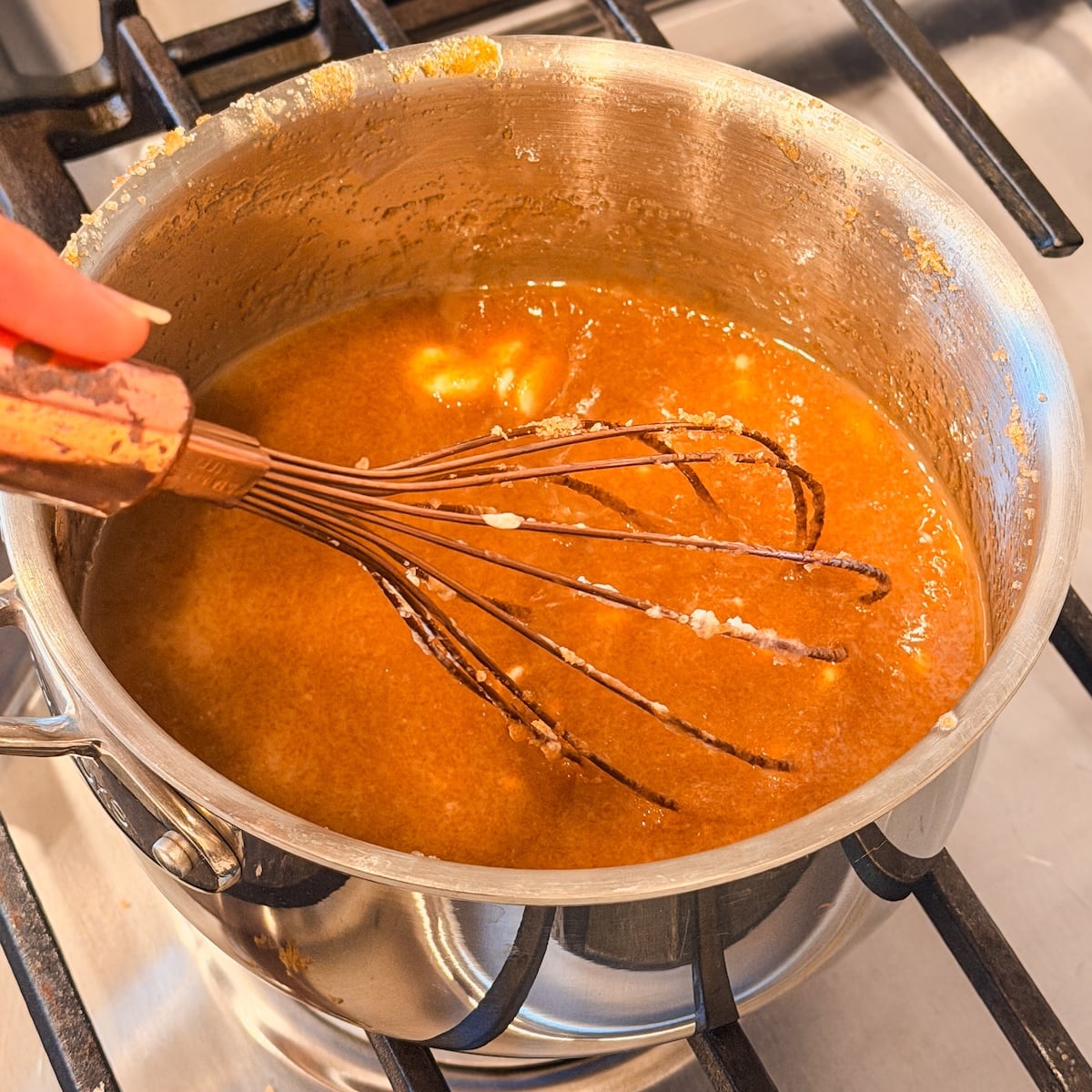 A hand uses a whisk to stir a golden-brown mixture in a saucepan on a stove, with melted butter and brown sugar
