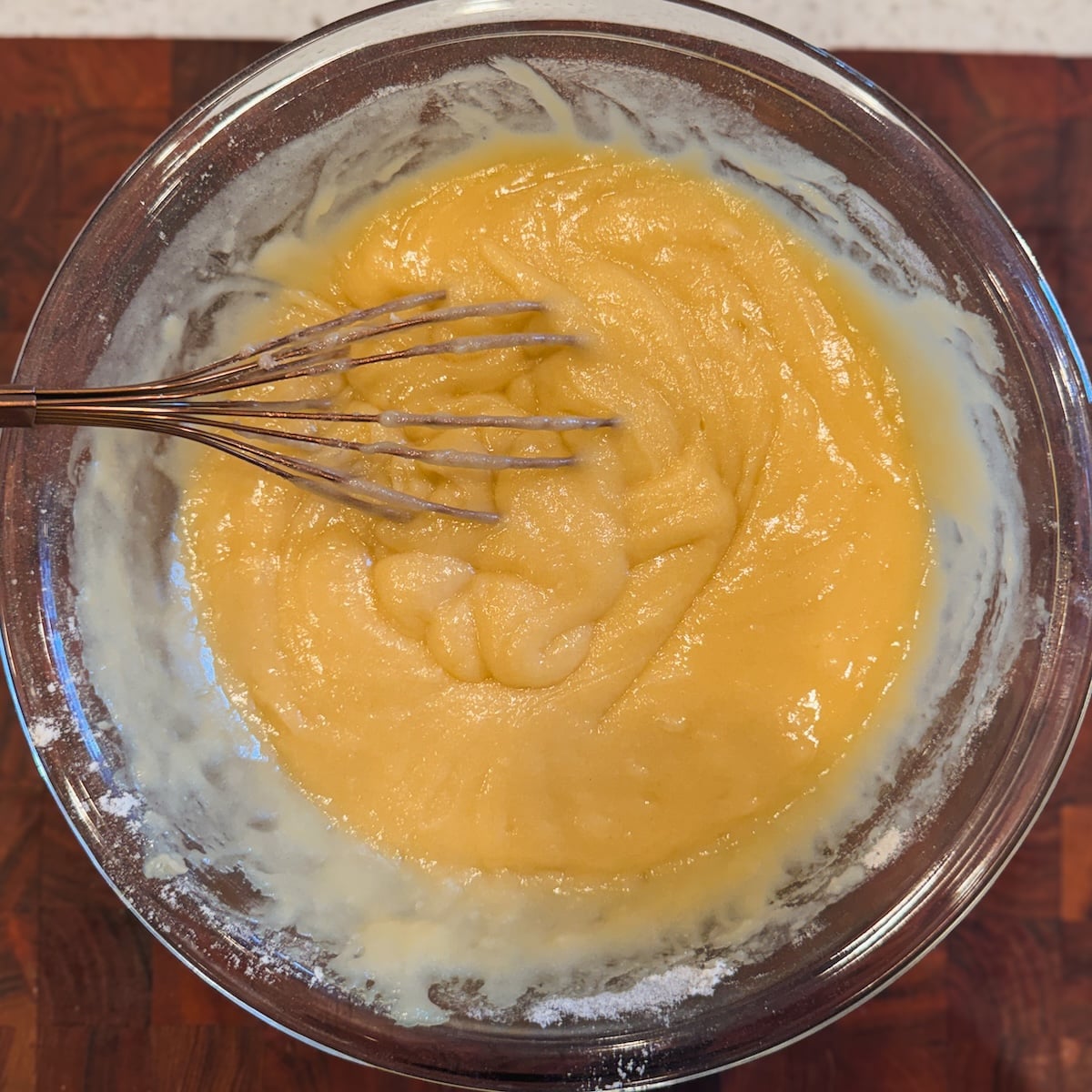 A glass bowl filled with thick cake batter being mixed with a metal whisk, sitting on a dark wooden surface.