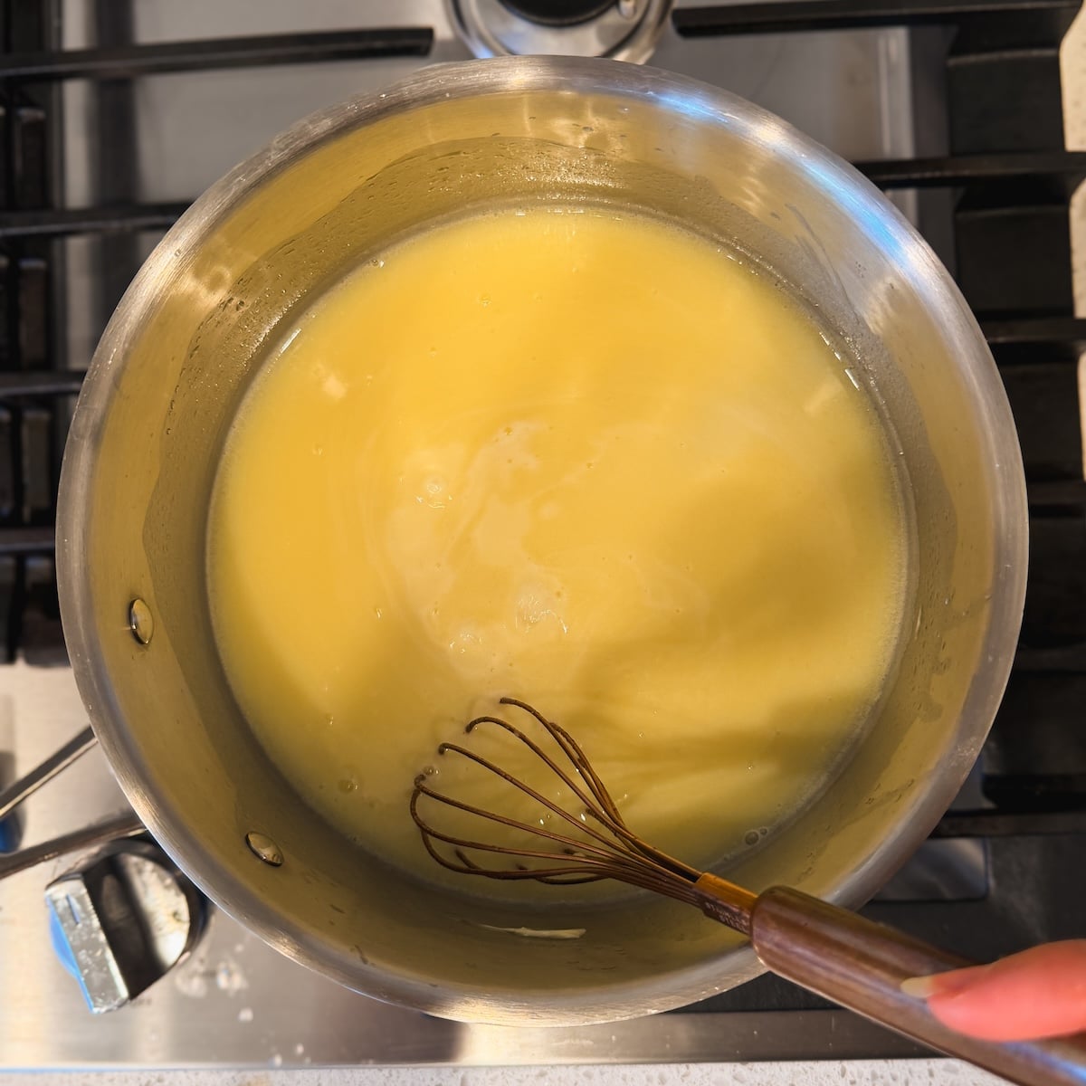 A hand whisking a thick yellow mixture in a stainless steel pot on a stovetop,