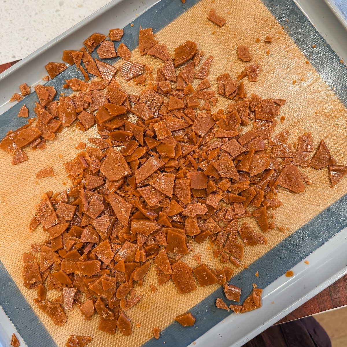 Shattered pieces of toffee candy spread out on a silicone baking mat and baking tray, with a white countertop visible in the background.