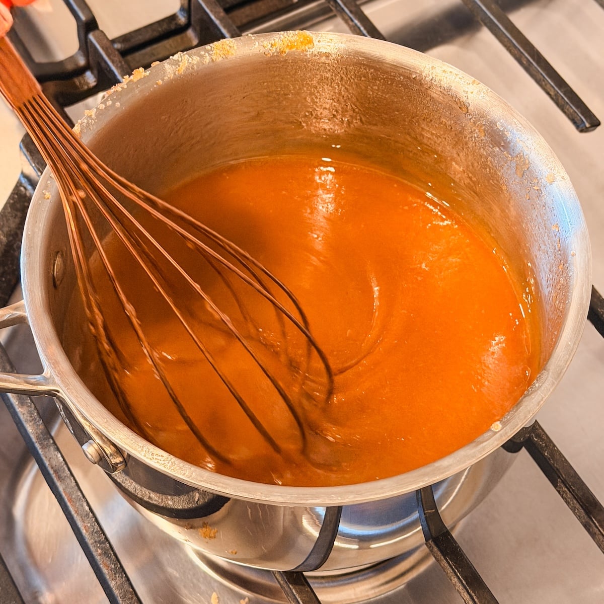 A whisk stirs a thick, orange-brown butterscotch sauce in a metal saucepan on a gas stove.