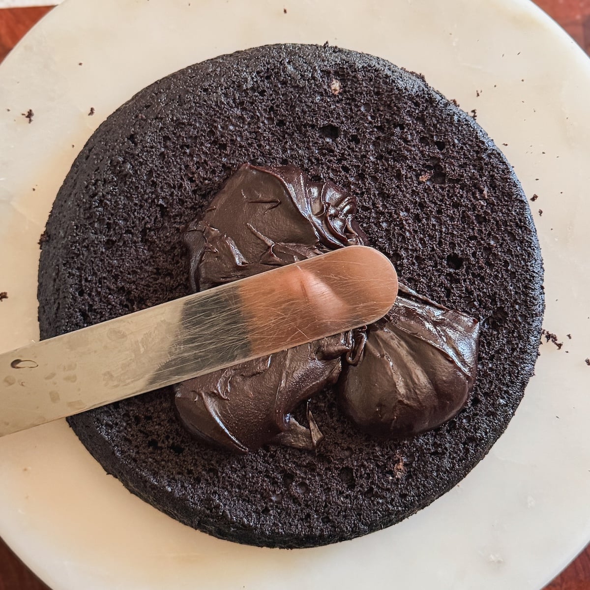 A metal spatula spreads chocolate fudge frosting on a round, dark chocolate cake layer placed on a white surface.