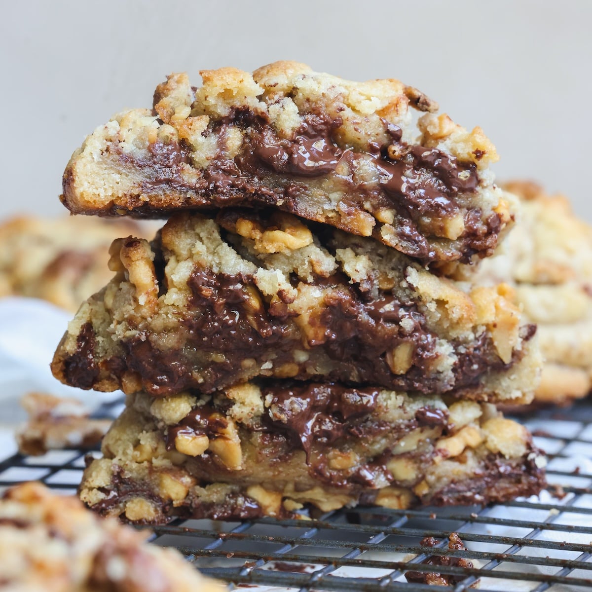 A stack of three thick chocolate chip and walnut cookies on a wire cooling rack. They look crispy outside and very gooey inside with melted chocolate puddles