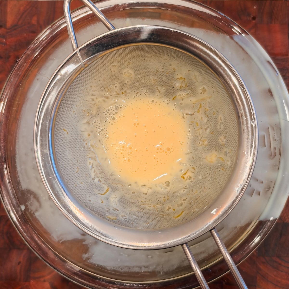 A metal sieve holding a yellow liquid is placed over a glass bowl, which sits on a wooden surface. The liquid appears to be strained, with some residue left in the sieve.