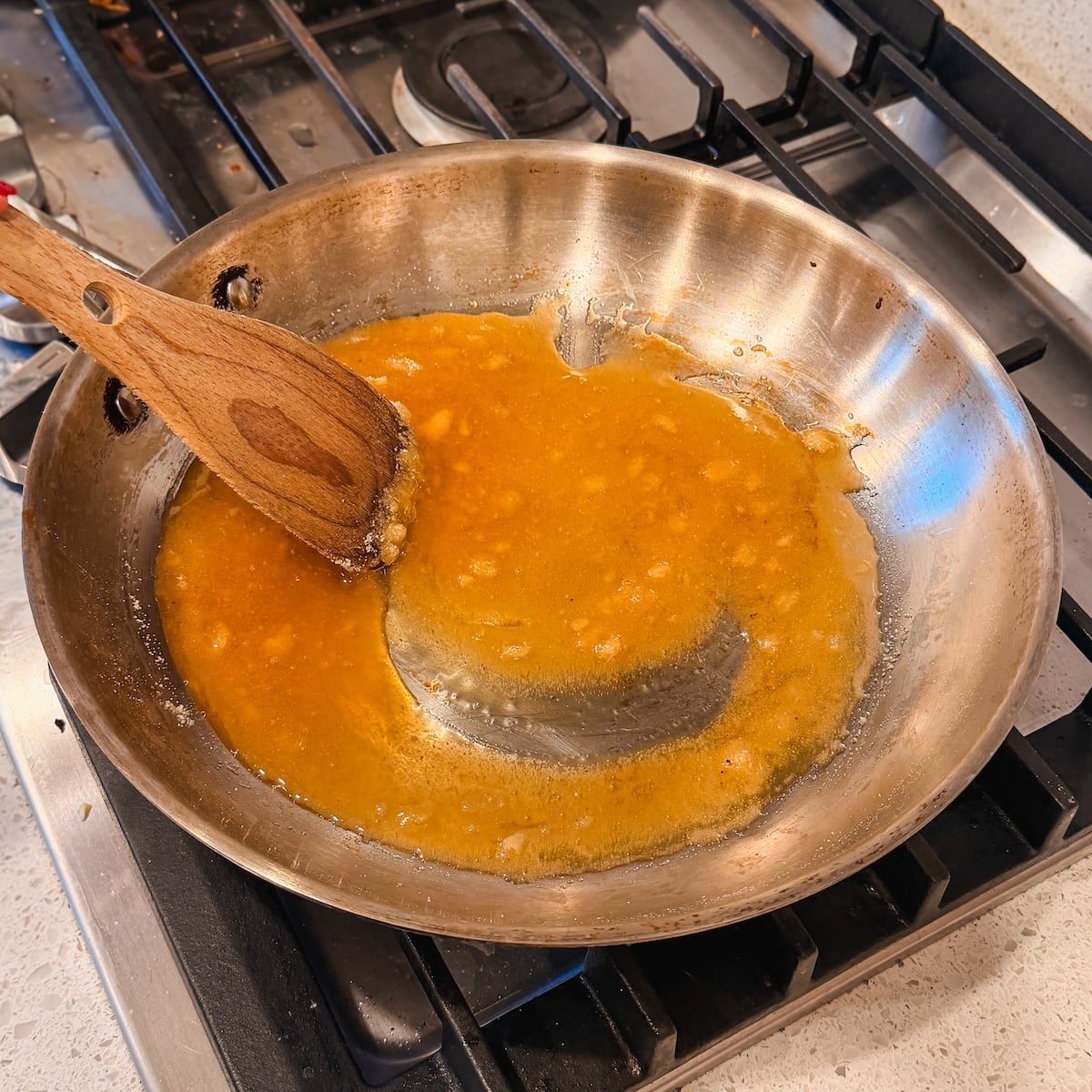 A wooden spatula stirs melting sugar in a stainless steel pan on a gas stove, with bits of sugar lumps visible in the sauce.
