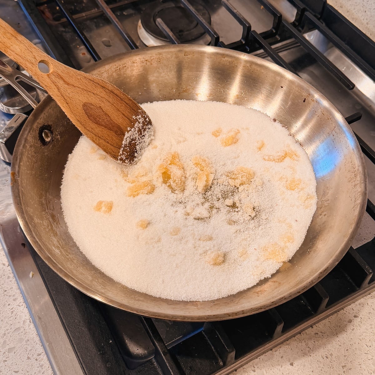 A stainless steel pan on a stove contains white sugar being stirred with a wooden spatula.