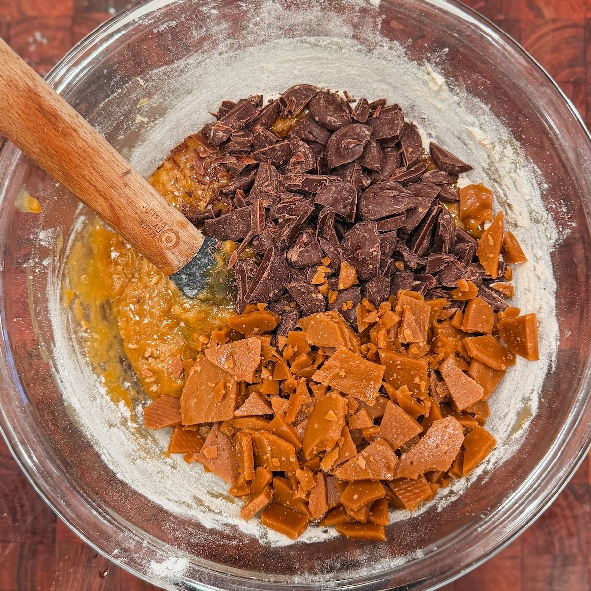 A clear bowl with cookie dough, dark chocolate chunks, and toffee pieces, shown from above. A wooden spoon rests inside the bowl, which is set on a reddish-brown surface.