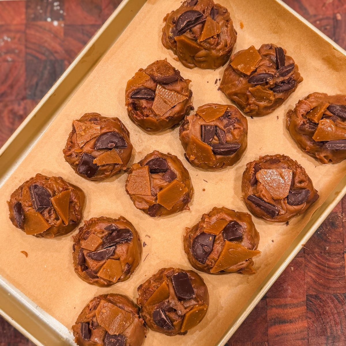 A baking tray lined with parchment paper holds twelve unbaked chocolate chip cookies topped with chocolate chunks and toffee pieces. The tray is on a wooden surface.
