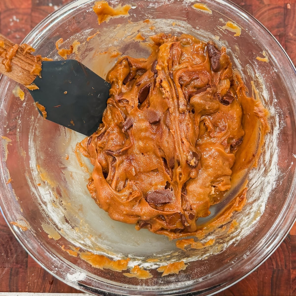 A glass bowl filled with toffee chocolate chip cookie dough being mixed with a black spatula on a wooden surface.