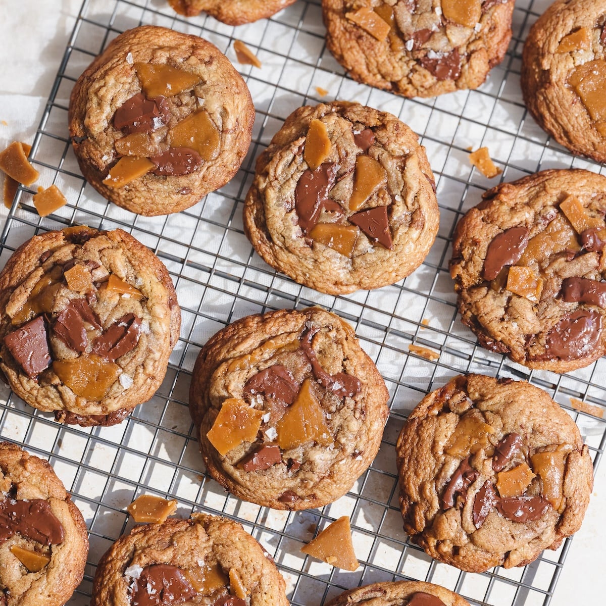 toffee chocolate chip cookies with toffee shards are cooling on a metal wire rack over parchment paper. The cookies are golden brown and topped with chocolate chunks and pieces of toffee.