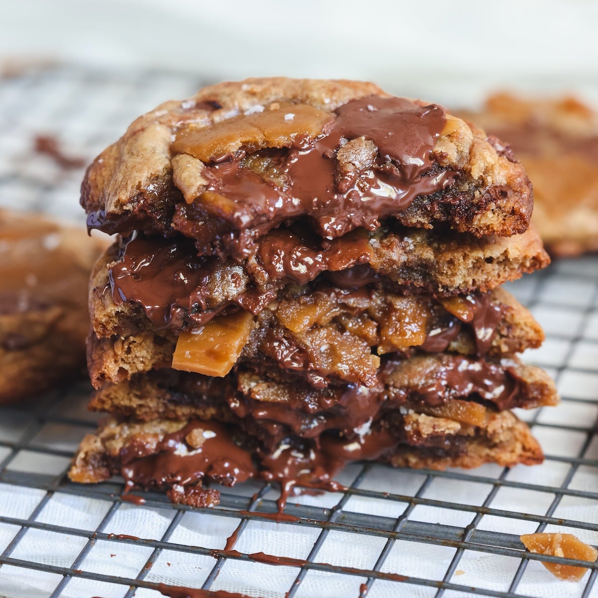 A stack of gooey chocolate chip cookies with melted chocolate and toffee bits, broken in half to show the rich, soft interior, sits on a cooling rack.