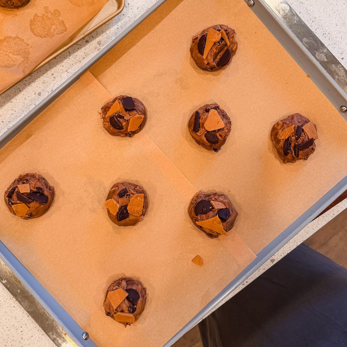 A baking tray with eight unbaked toffee chocolate chip cookie dough balls on parchment paper, ready to be baked.