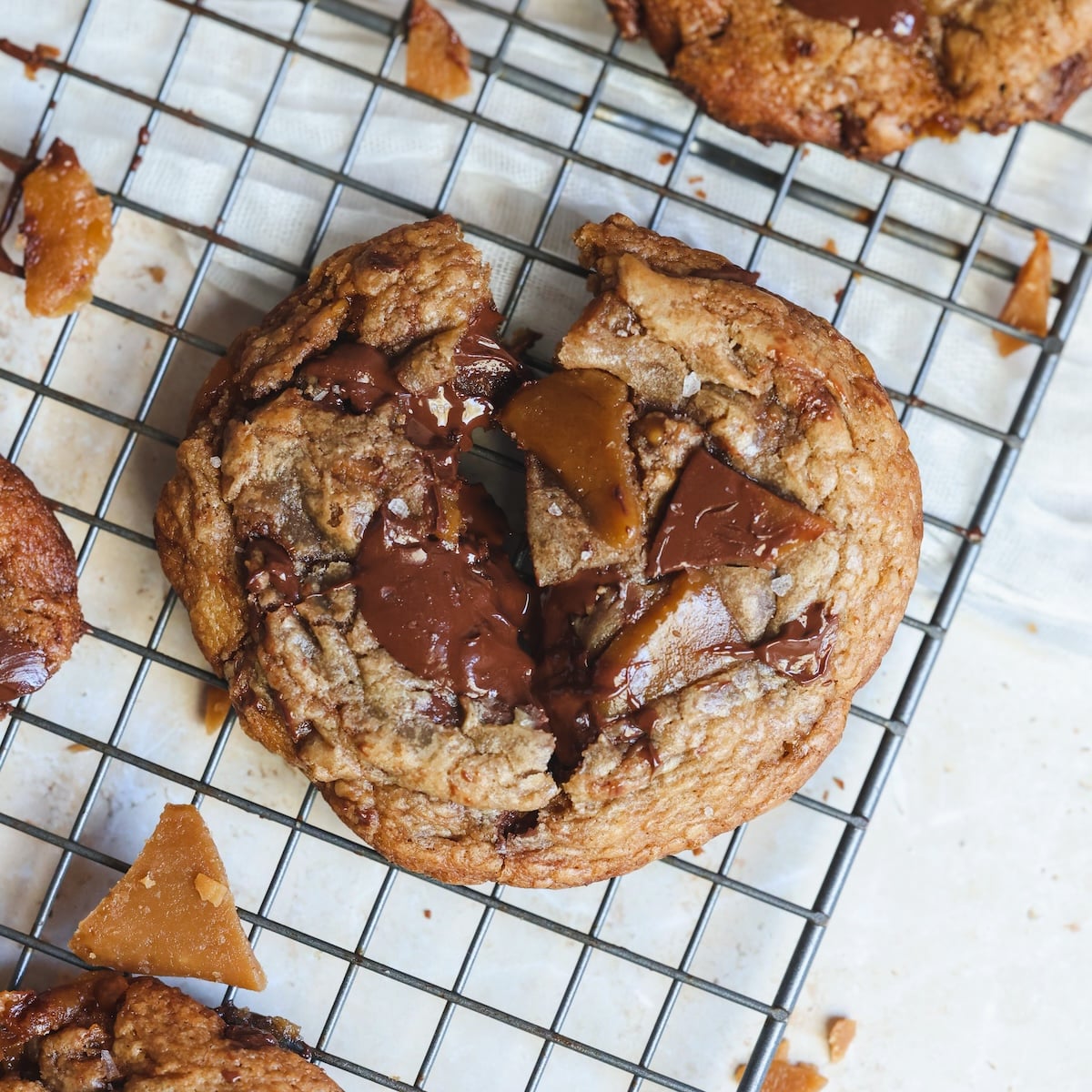 A chocolate chip cookie with toffee chunks is broken in half on a cooling rack, with melted chocolate and crumbs visible around it.