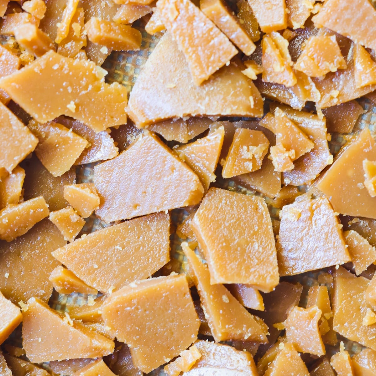 Broken pieces of golden brown toffee scattered on a light-colored surface, showing a brittle, sugary texture with sharp edges and some surface shine.